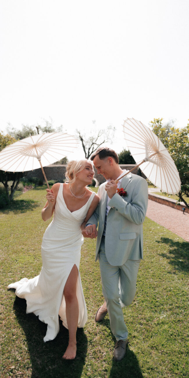 Happy bride and groom walking on a meadow with umbrellas during a luxury destination wedding in Europe.